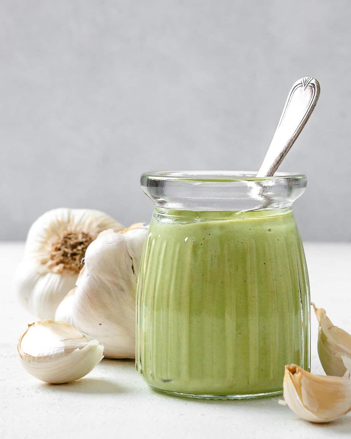 Cremige Vegane Knoblauchsoße 2 Fertige cremige vegane Knoblauchsauce in einem Glasgefäß mit Knoblauch im Hintergrund