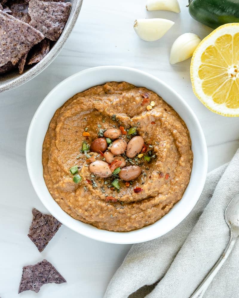 Gewürzter Bohnen-Dip 2 completed Spiced Refried Bean Dip in a bowl against a white background with ingredients spread in the back