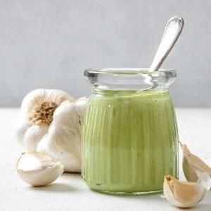 Cremige Vegane Knoblauchsoße 11 Fertige cremige vegane Knoblauchsauce in einem Glasgefäß mit Knoblauch im Hintergrund