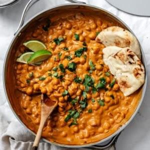 Curry-Kichererbsen 12 Kichererbsen-Curry in einer großen Pfanne mit einem Holzlöffel und veganem Naan-Brot daneben.