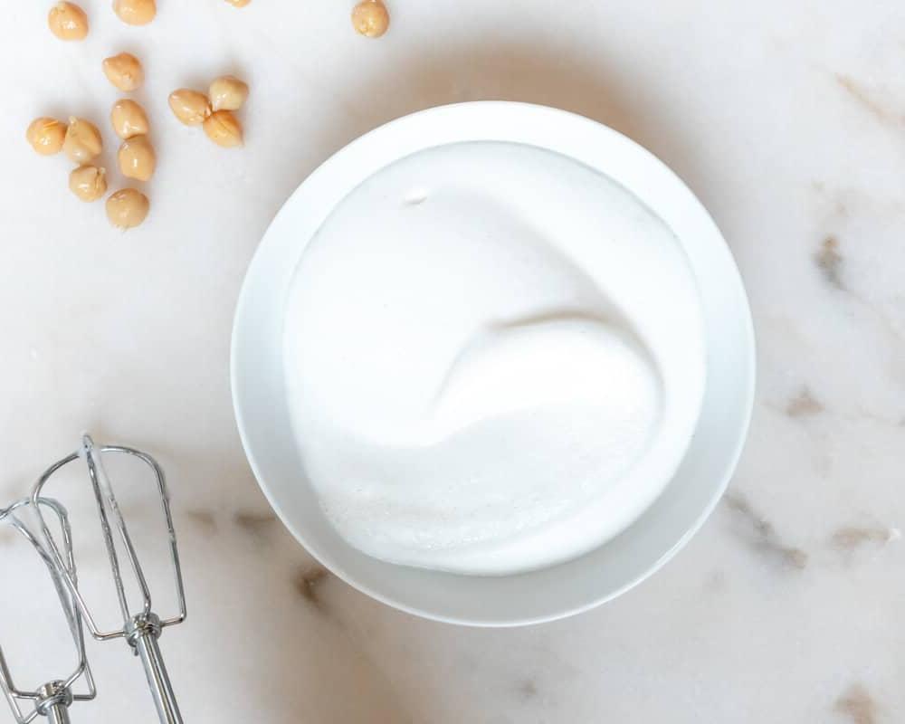 Eine Schale mit fluffigem Kichererbsenwasser oder Aquafaba mit einigen Kichererbsen daneben