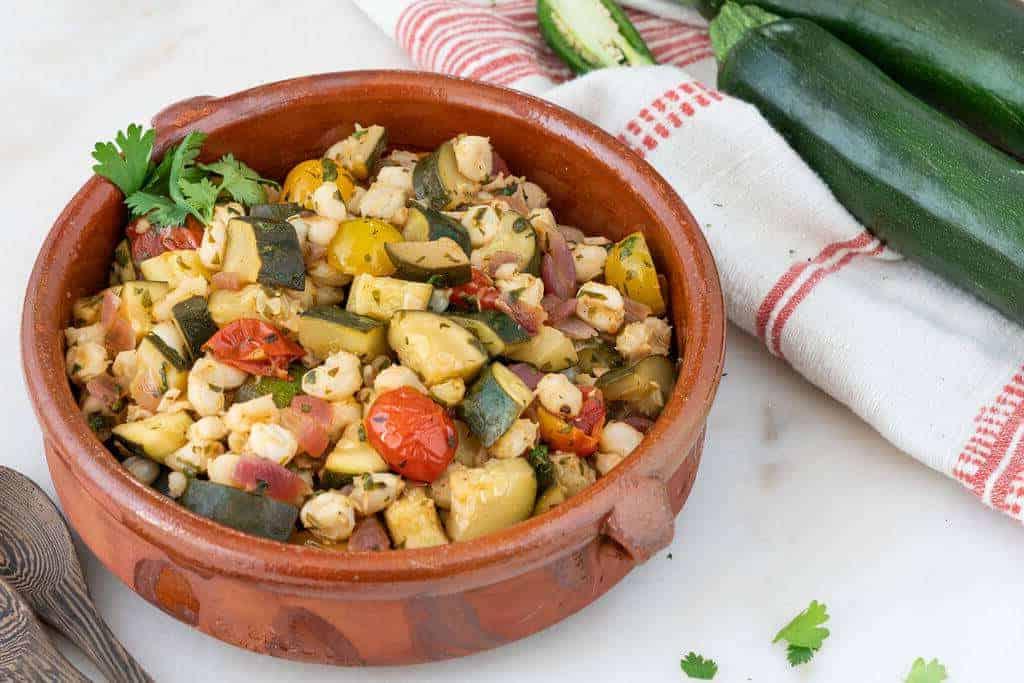 Braune Schale mit Zucchini und Hominy (Mais) mit einem rot-weißen Tuch auf weißem Hintergrund