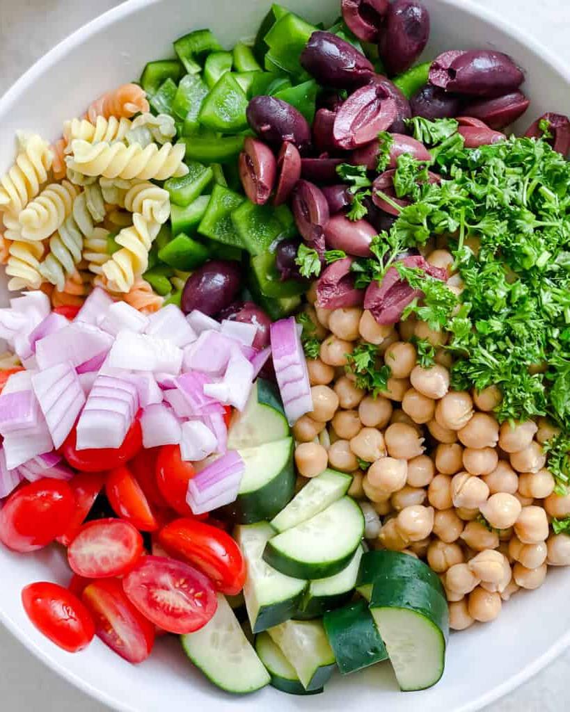 **Mediterraner veganer Nudelsalat** 5 Verfahrensschritt, der das Hinzufügen von Zutaten zu einer Schüssel zeigt
