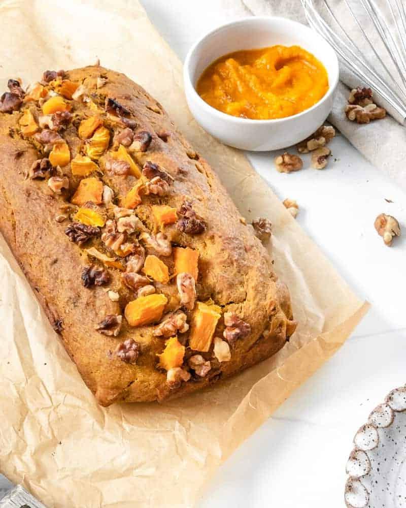 Veganes Kürbisbrot 7 Fertiger Kürbisbrotlaib auf Backpapier mit Zutaten im Hintergrund