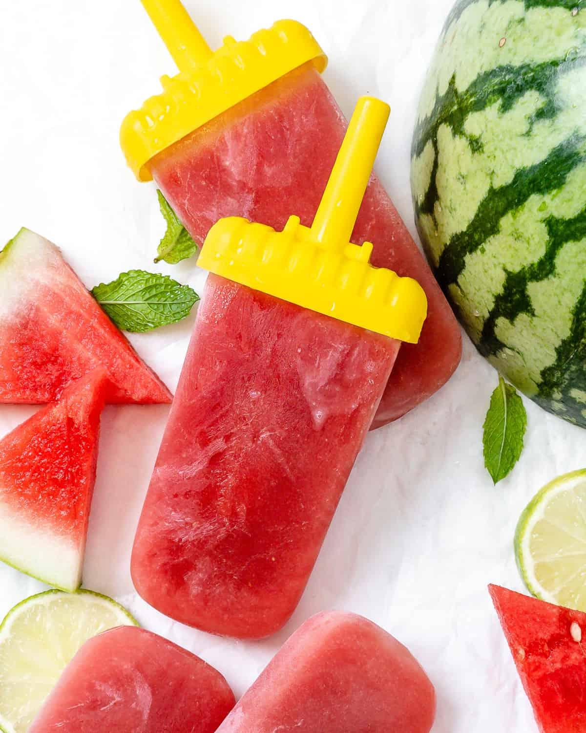 Gesundes Wassermelonen-Stieleis 7 Zwei fertige Wassermelonen-Eis am Stiel mit Wassermelonenstücken im Hintergrund