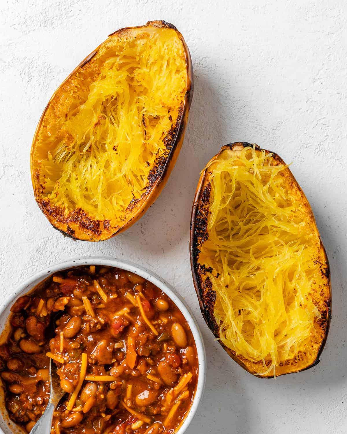 Chili-Käse-gefüllter Spaghettikürbis 8 two halves of spaghetti squash alongside a bowl of chili against white background