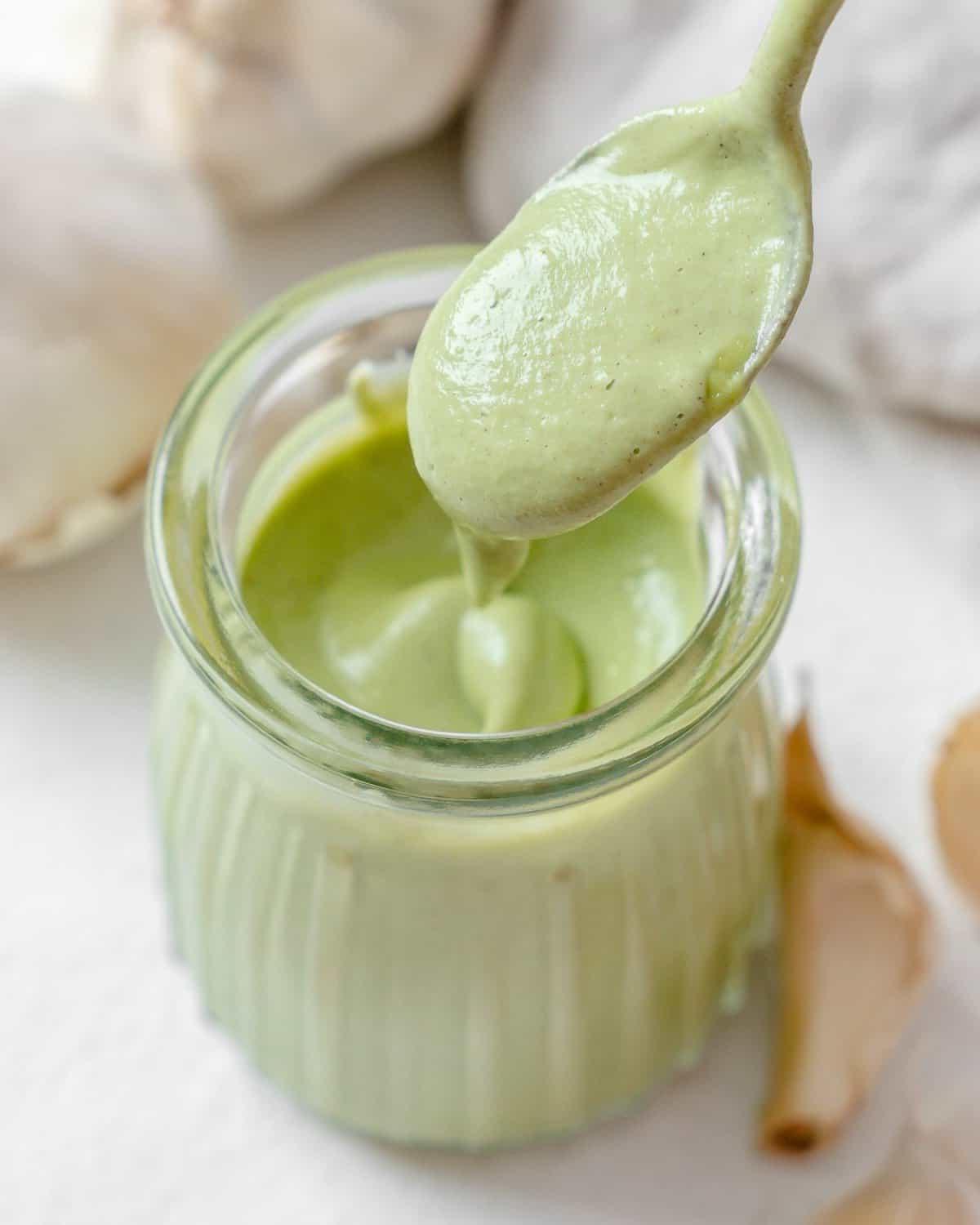 Cremige Vegane Knoblauchsoße 9 Fertige cremige vegane Knoblauchsauce in einem Glasgefäß, ein Löffel zeigt die Konsistenz, mit Knoblauch im Hintergrund