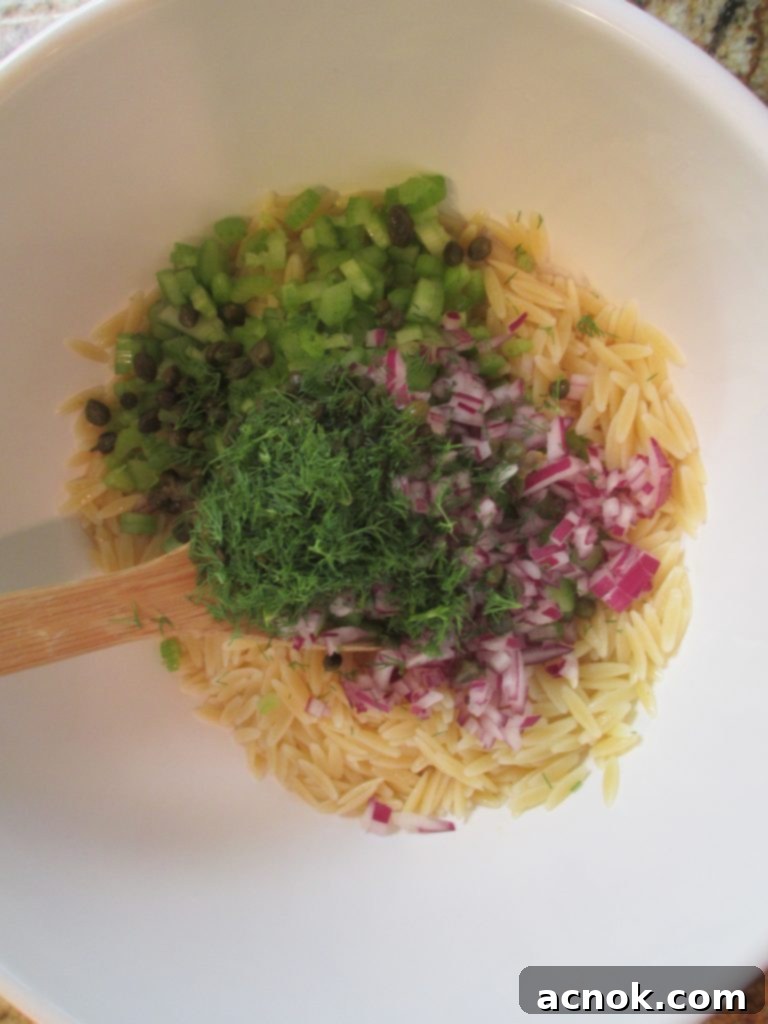 All salad ingredients – orzo, vegetables, dill, and capers – combined in a bowl before adding salmon and dressing.