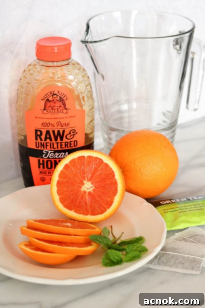 Ingredients for iced orange honey green tea, including tea bags, fresh oranges, mint leaves, and a jar of honey, laid out on a wooden surface.