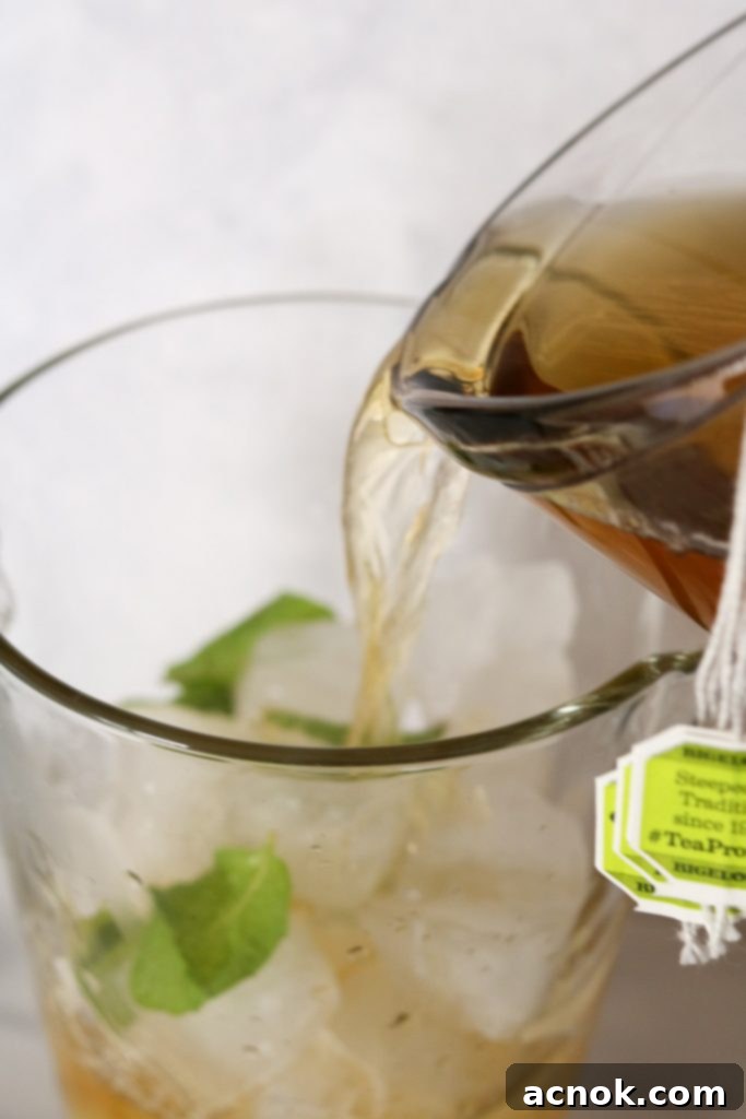 A hand pouring warm green tea from a glass measuring cup over fresh mint leaves and ice cubes in a glass pitcher.