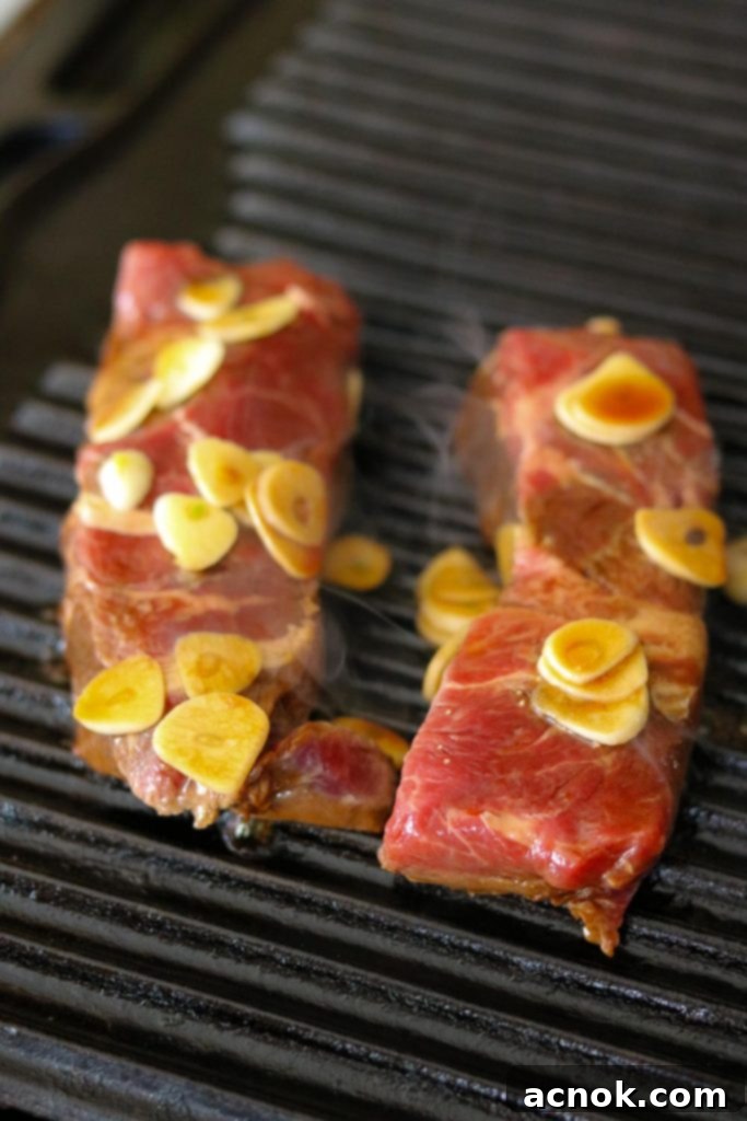 Raw flat iron steaks and garlic slices placed on a hot stovetop grill pan.
