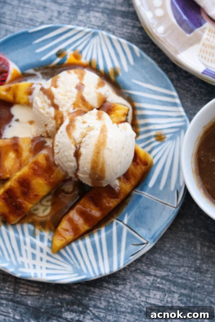 Two scoops of vanilla ice cream with grilled pineapple spears and generous spiced rum butter sauce on a white plate, garnished with mint.