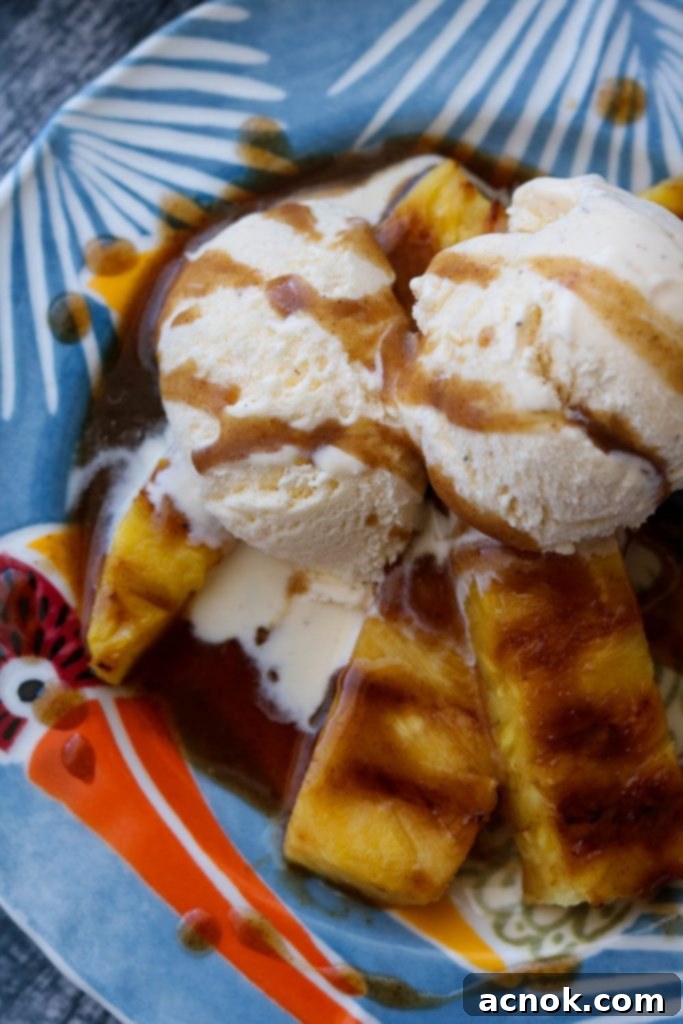 A plated dessert featuring grilled pineapple, two scoops of vanilla ice cream, and a generous drizzle of spiced rum butter sauce.