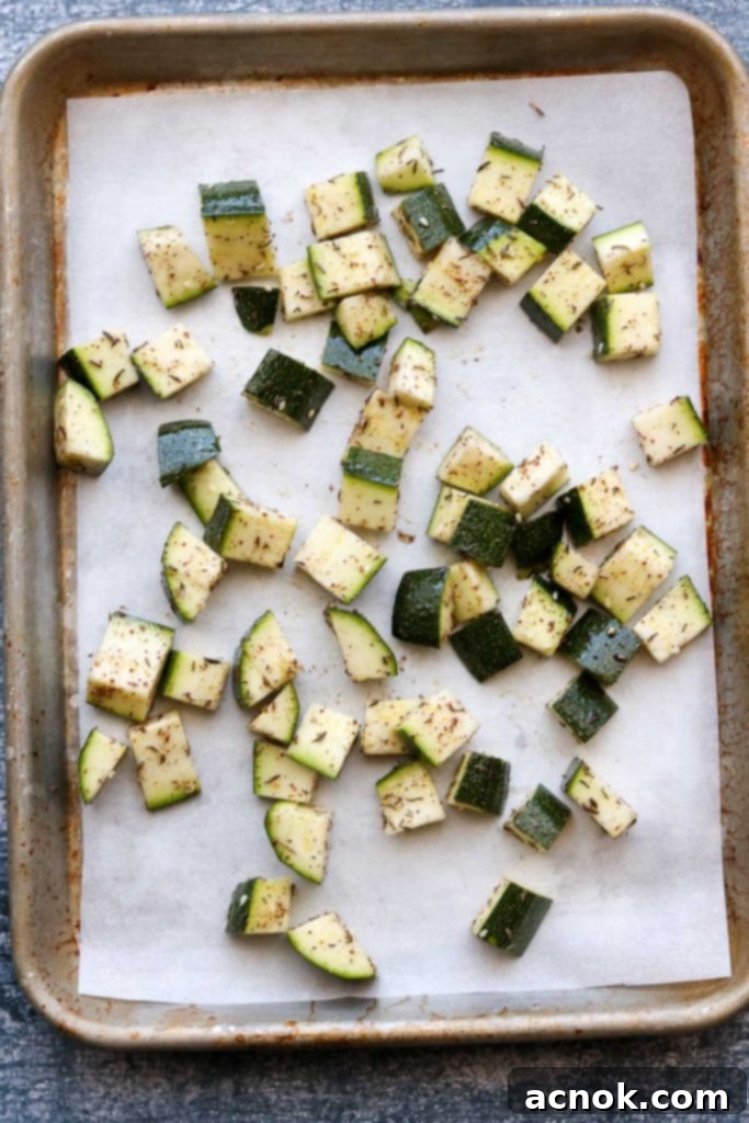 Seasoned zucchini cubes spread in a single, uncrowded layer on a baking sheet lined with parchment paper, ready for the oven