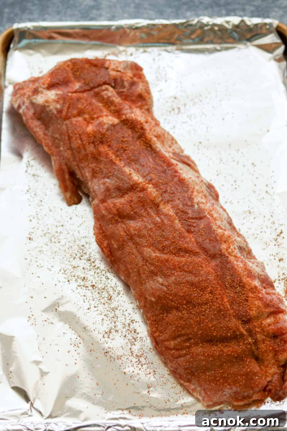 Raw ribs with a rich dry rub on a half sheet pan, covered securely with aluminum foil, resting before cooking.
