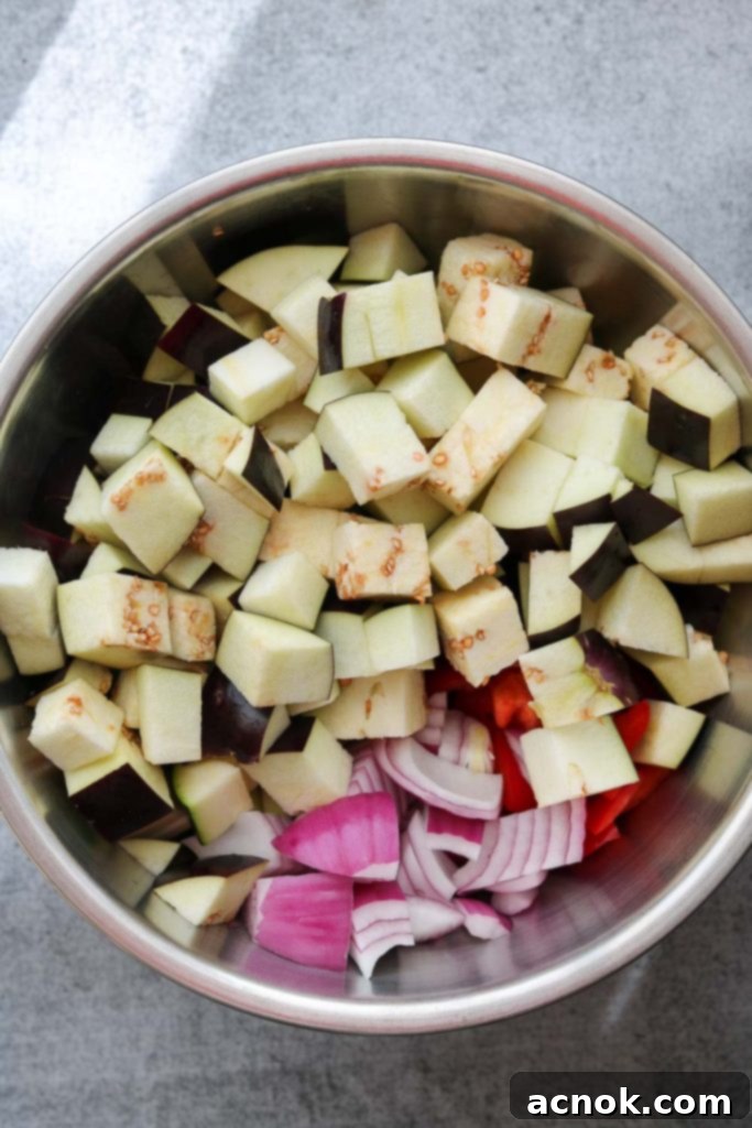 Cubed eggplant, zucchini, red bell pepper, and red onion in a large white bowl, ready for seasoning.