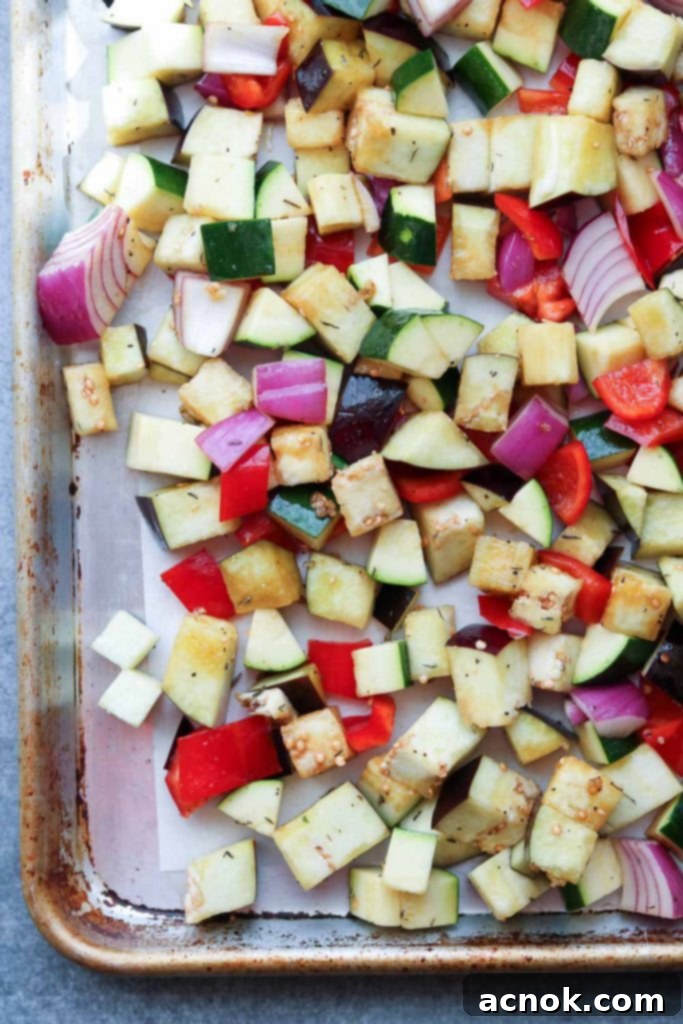 Marinated cubed vegetables spread in a single layer on a parchment-lined rimmed baking sheet, ready for roasting.