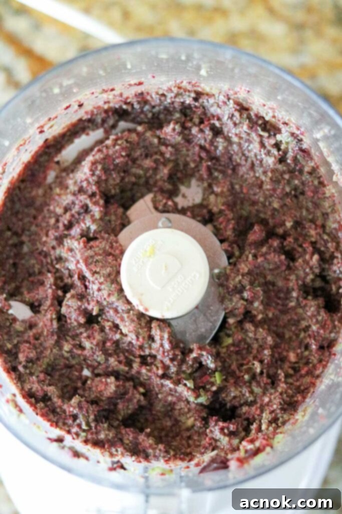 Olive oil being slowly poured through the feed tube of a food processor while pulsing, creating a creamy tapenade texture.