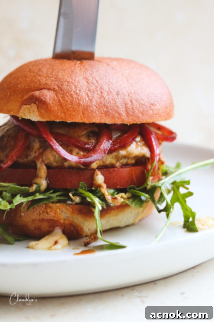 Delicious Pesto Parmesan Chicken Burger with fresh arugula, tomato, and marinated red onions on a gluten-free bun, served on a white plate next to a knife.