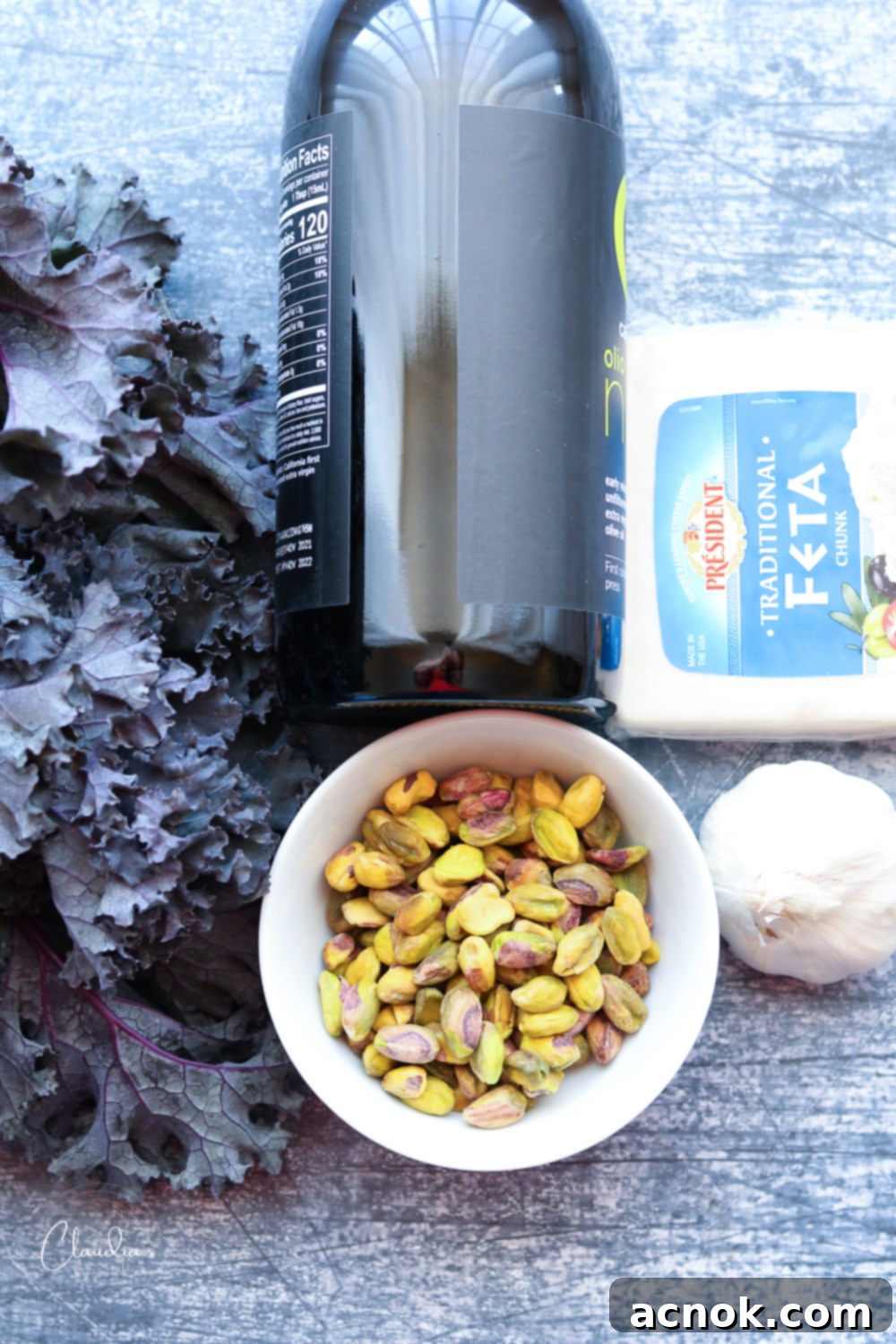 A vibrant flat lay of fresh ingredients for kale pistachio feta pesto: purple kale, a bottle of extra virgin olive oil, garlic cloves, raw pistachios, a block of feta cheese, lemon, and seasonings.