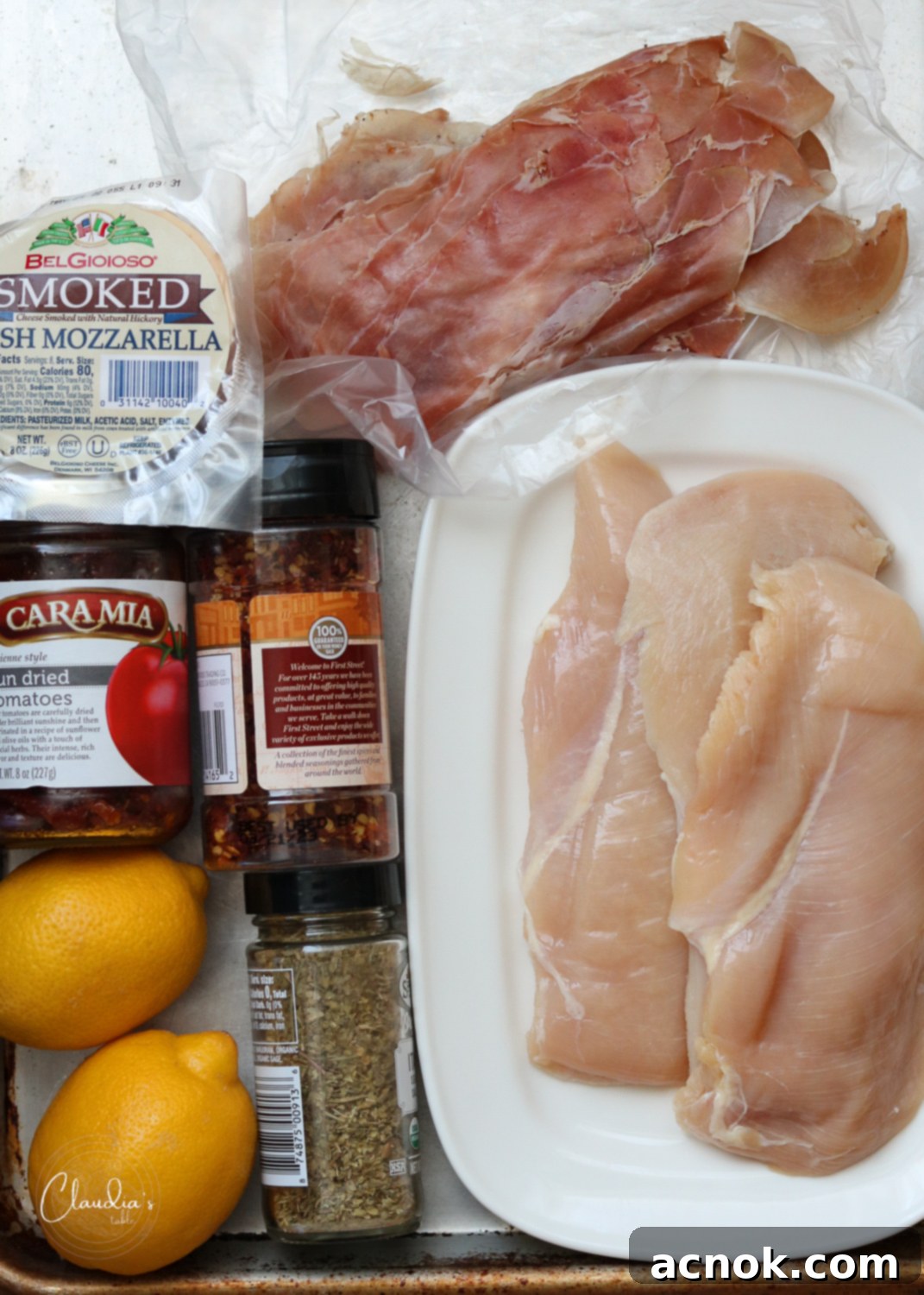 Ingredients for grilled chicken involtini laid out on a baking sheet: chicken, prosciutto, sun-dried tomatoes, smoked mozzarella, and spices.