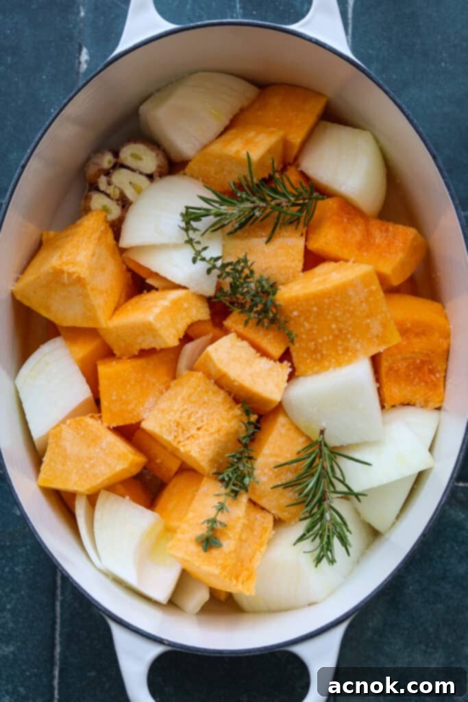 Large chunks of butternut squash and onions, whole garlic head with trimmed tops, fresh herbs, and olive oil in a Dutch oven.