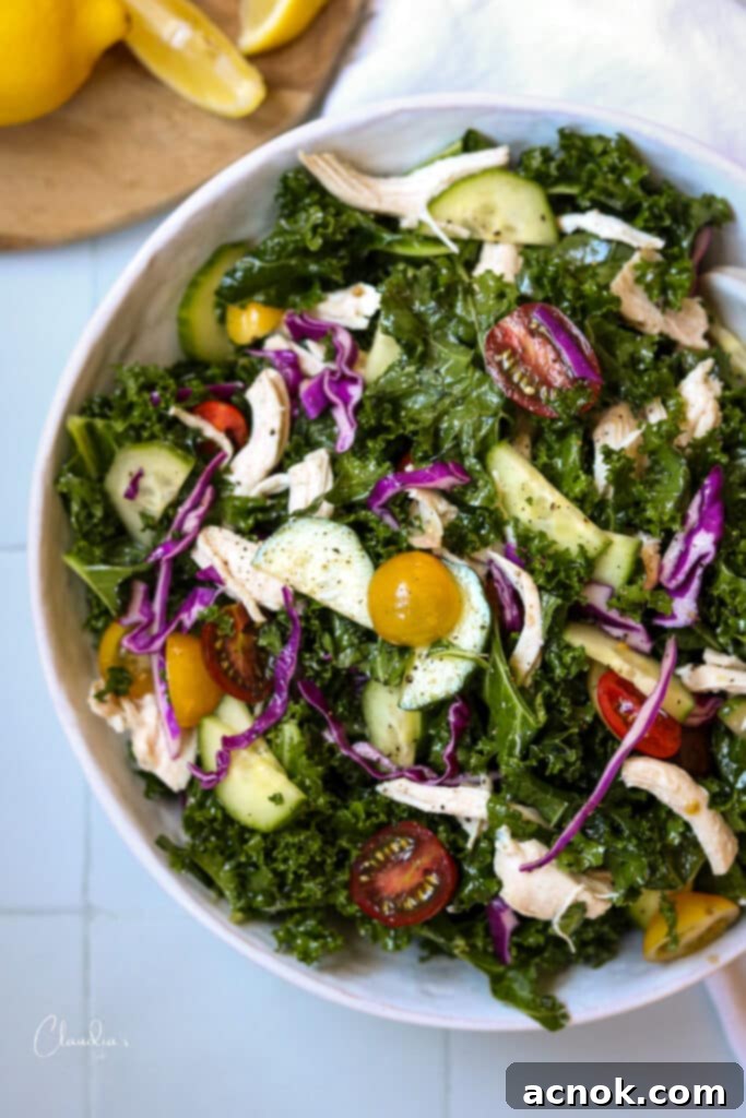 Vibrant Mediterranean kale salad in a white bowl on a white table, showcasing fresh ingredients like kale, chicken, cucumber, and cabbage.